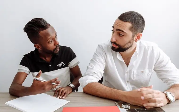 Two people talking over documents at a table, one holding a pen
